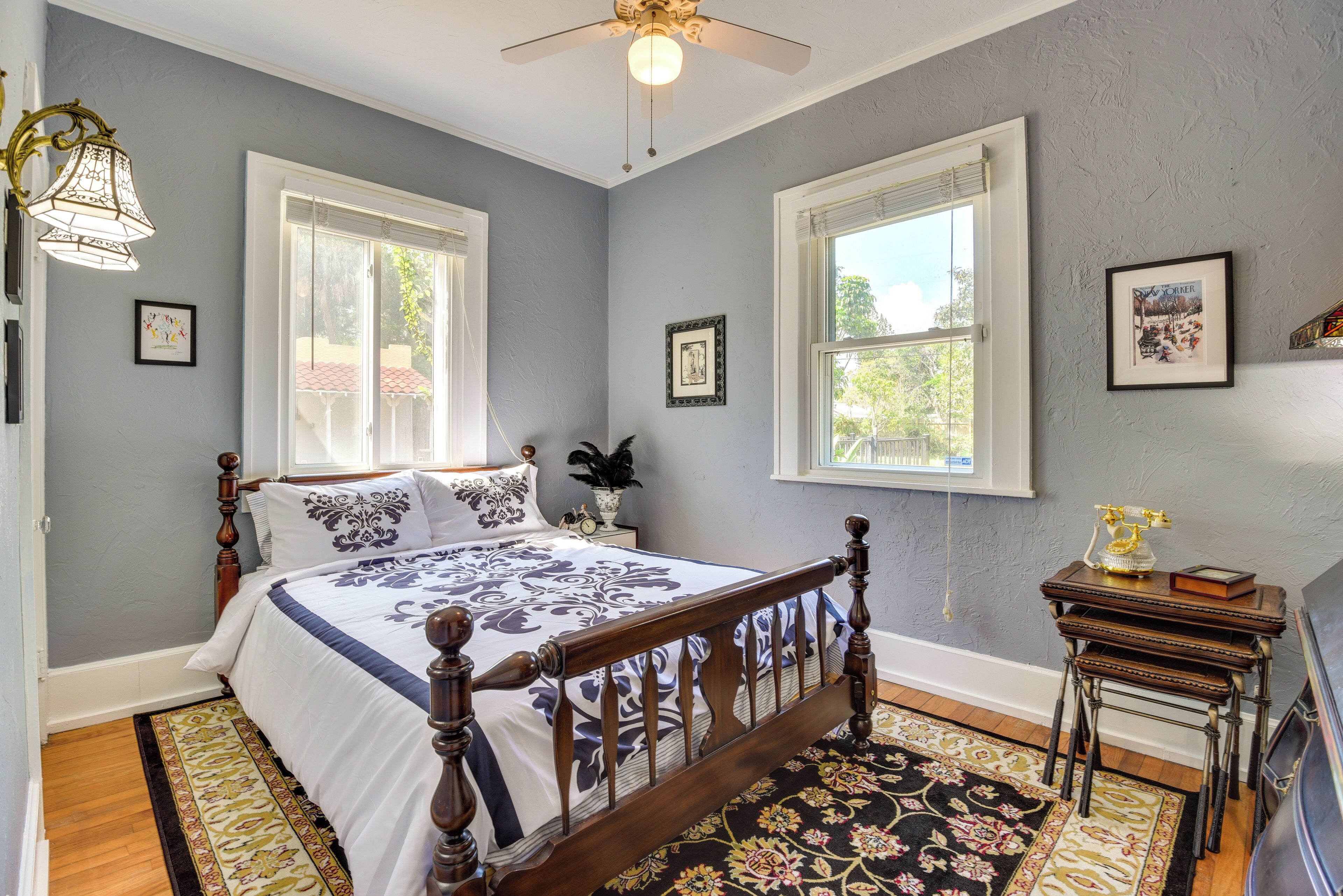 A bedroom with a wooden bed, two windows, a ceiling fan, and a small table with a lamp.