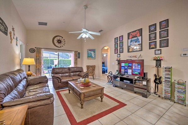 A living room with a brown leather sofa, a wooden coffee table, a ceiling fan, and a TV stand with a guitar and a DVD rack. - 10 Mi to Disney + Heated Pool: Florida Abode! (Clermont)