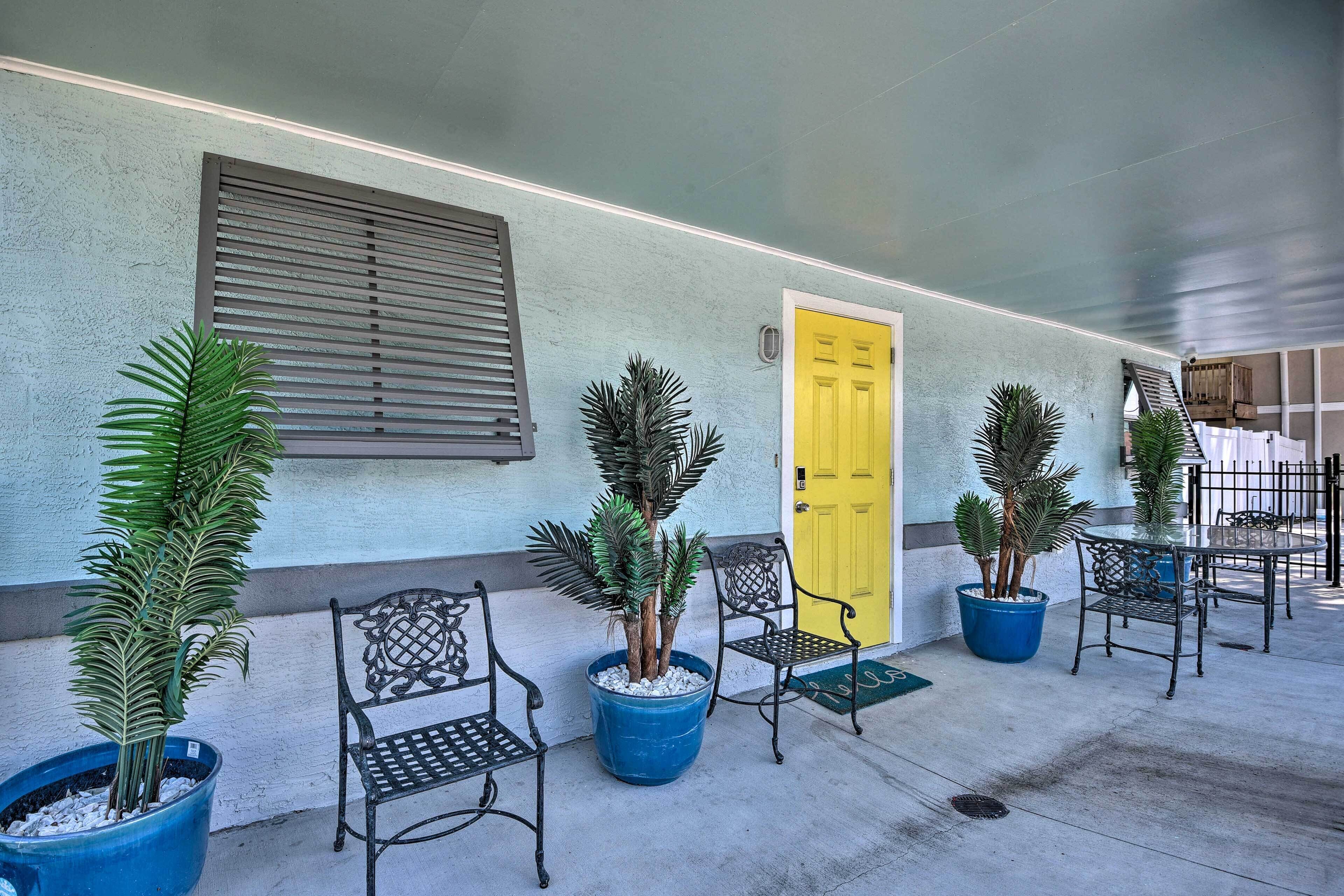 A covered outdoor seating area with potted plants and metal chairs, a yellow door, and a window with blinds.