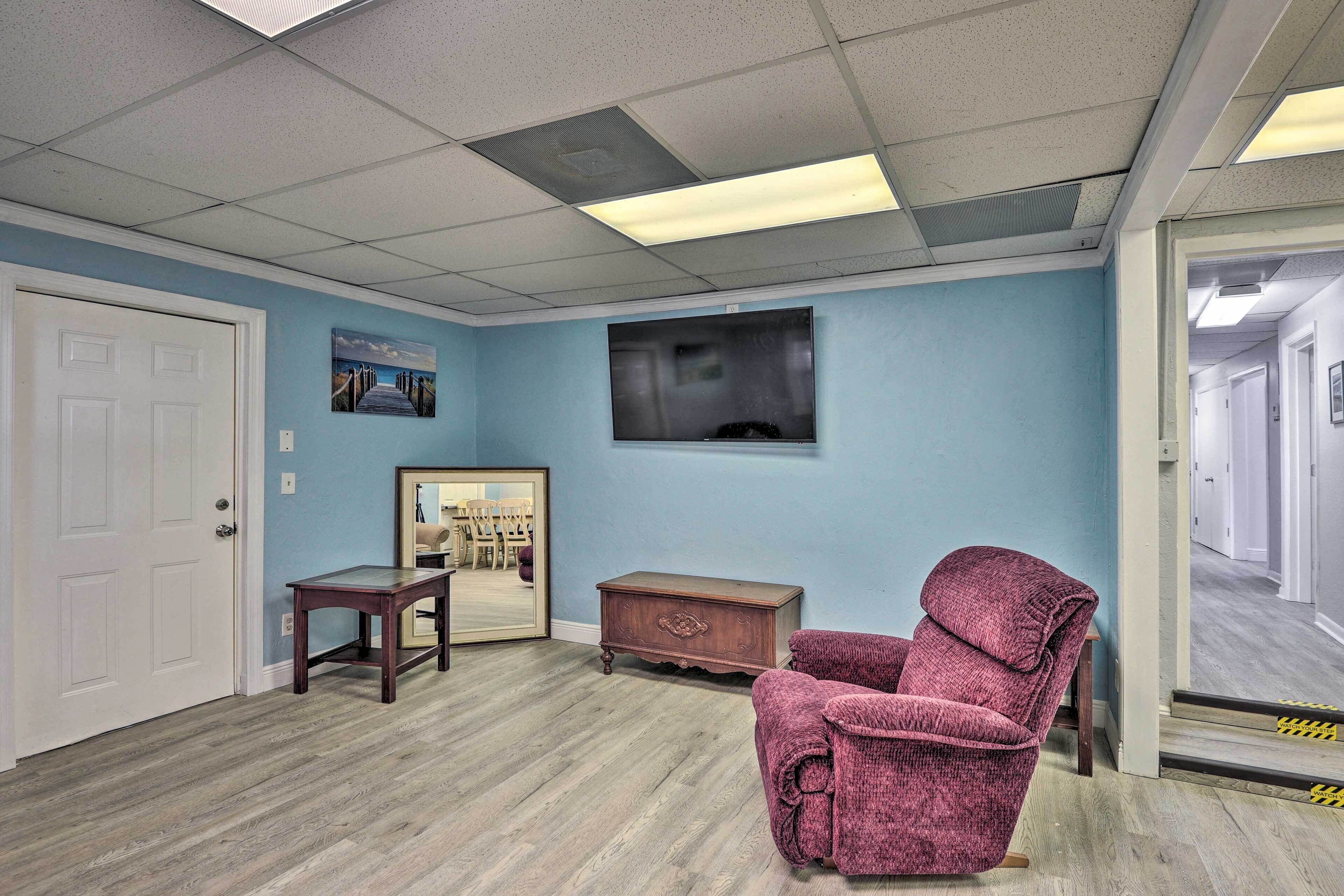 A room with a wooden floor, a door, a small table, a chair, a dresser, and a television mounted on the wall.