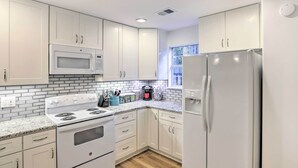 Une cuisine avec des meubles blancs, un four à micro-ondes, une cuisinière, un réfrigérateur et une fenêtre avec des stores.
