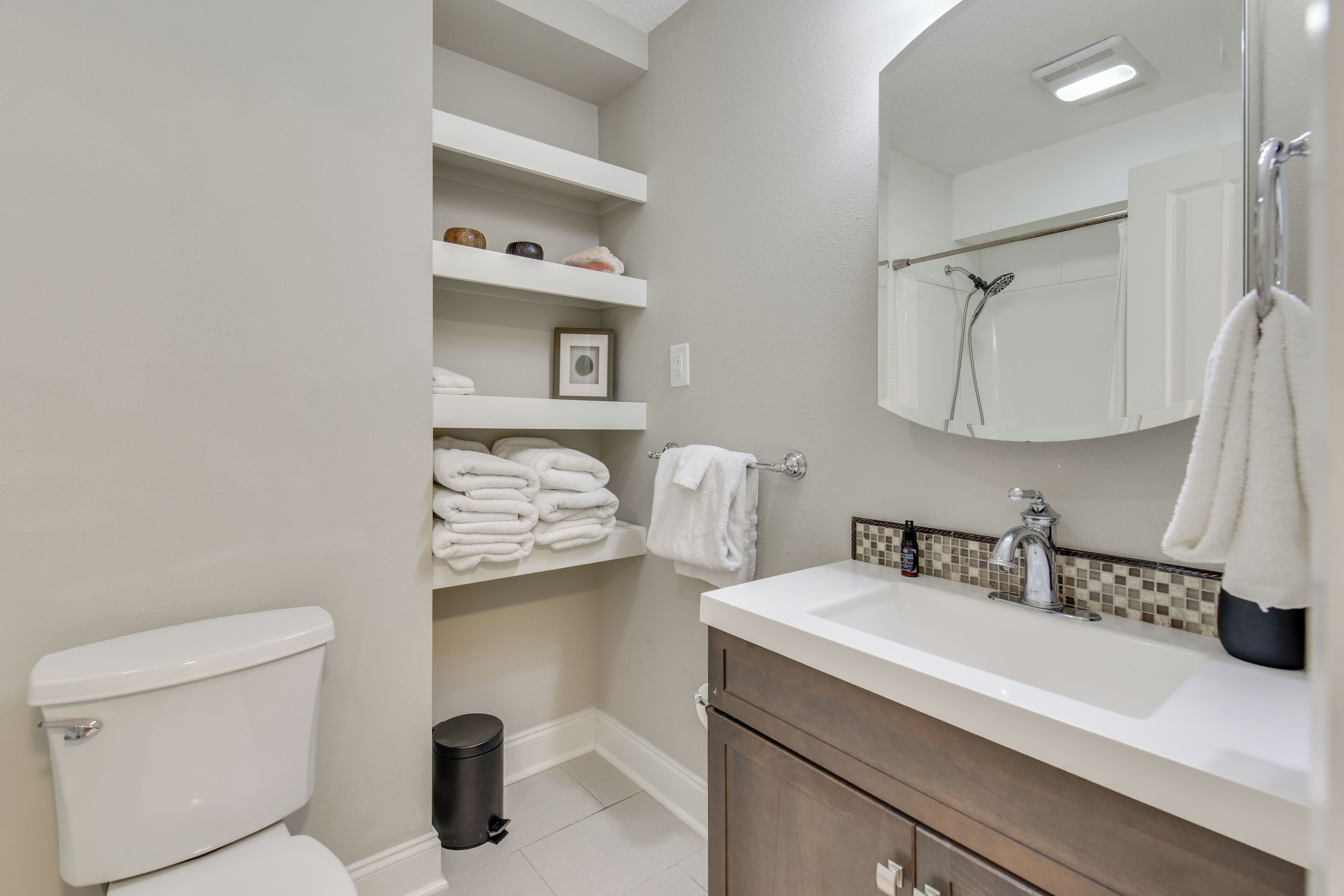 A modern bathroom with a toilet, sink, and mirror. Shelves with towels and toiletries are visible.