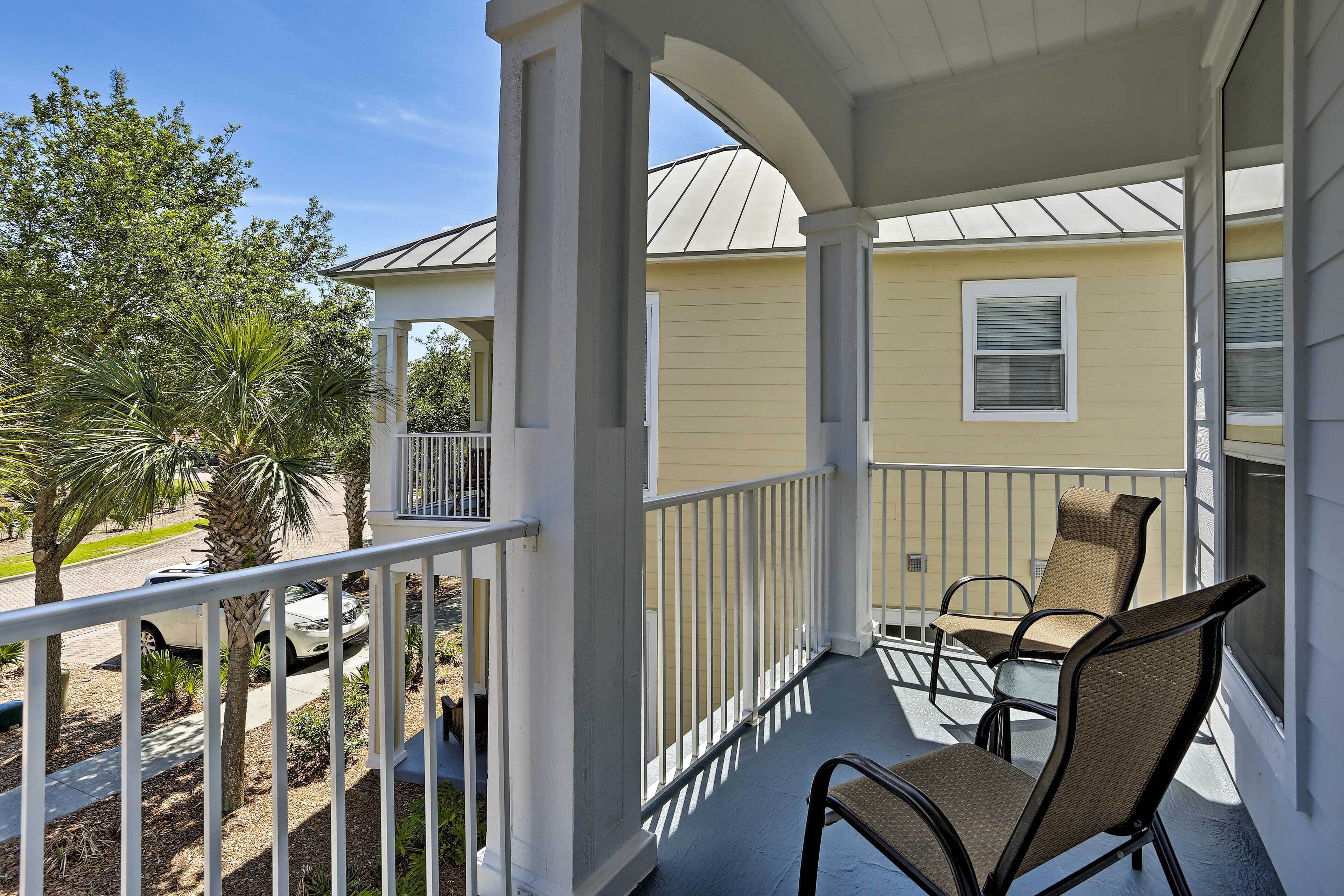 A covered porch with two wicker chairs, a white railing, and a view of a yellow house and palm trees.