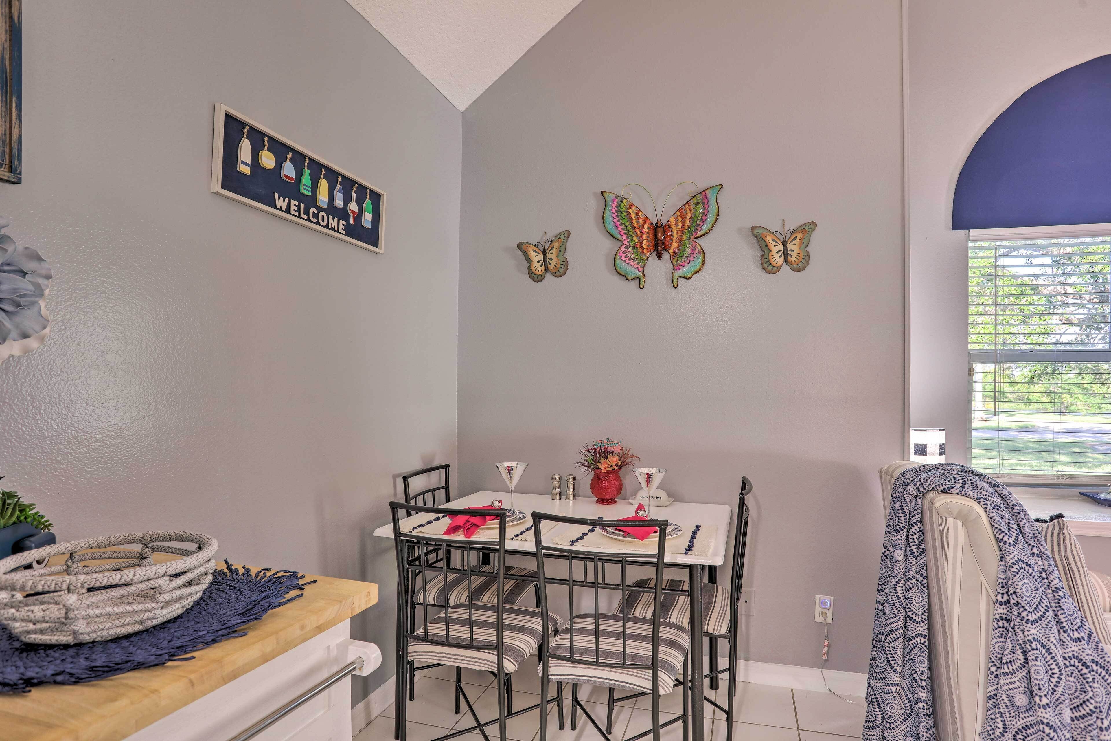 A dining area with a table set for four, chairs, a 'WELCOME' sign, decorative butterflies, and a window with blinds.