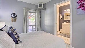 A bedroom with a bed, a bedside table, a wall-mounted TV, a window with curtains, and a bathroom visible through an open door. - Stuart Apartment w/ Patio Near St Lucie River! (Stuart)