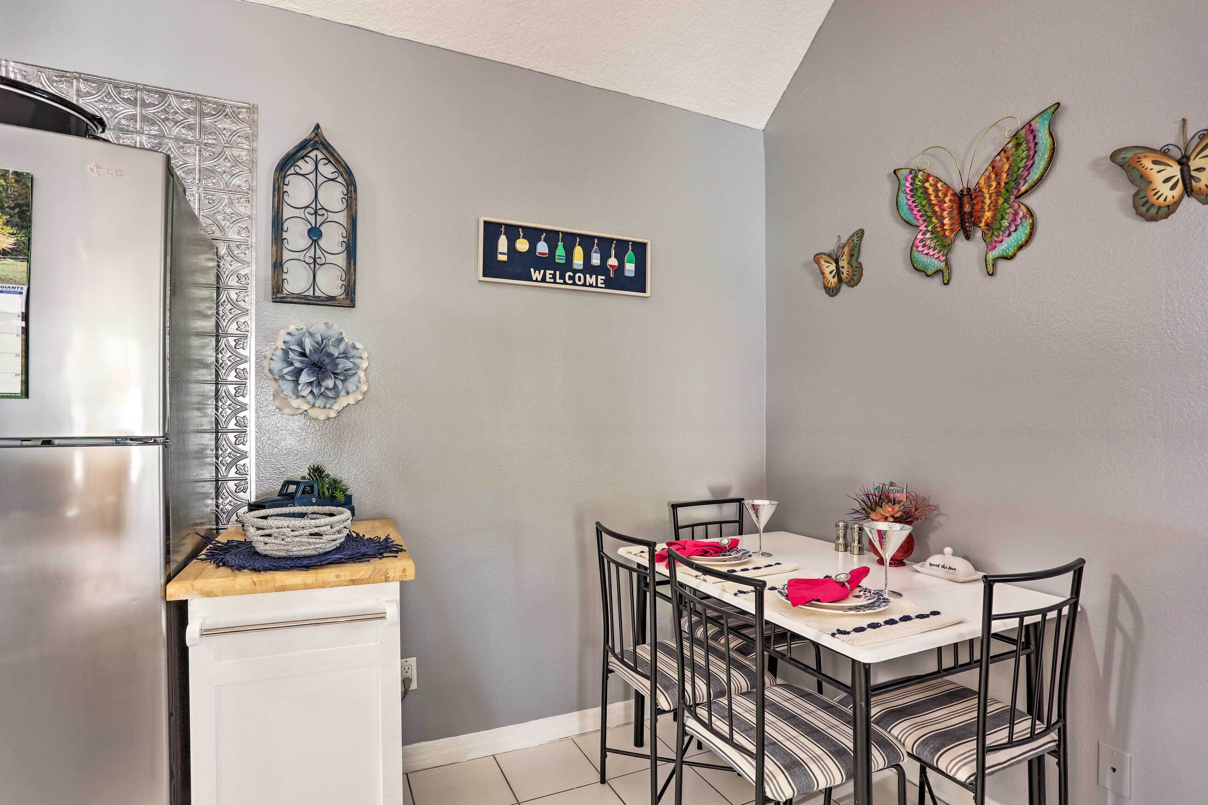 A dining area with a table set for two, a refrigerator, decorative wall art, and a welcome sign.