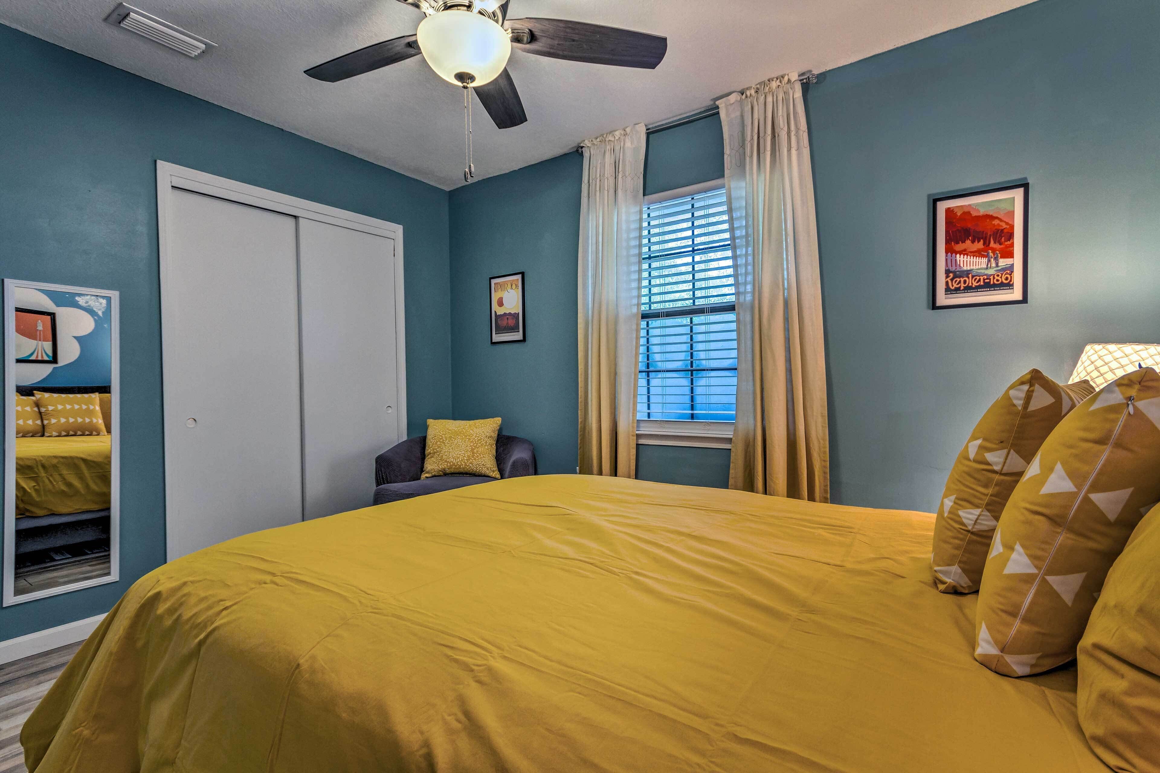 Une chambre à coucher avec un lit, un ventilateur de plafond, une fenêtre avec des rideaux, une porte et un tableau encadré accroché au mur.