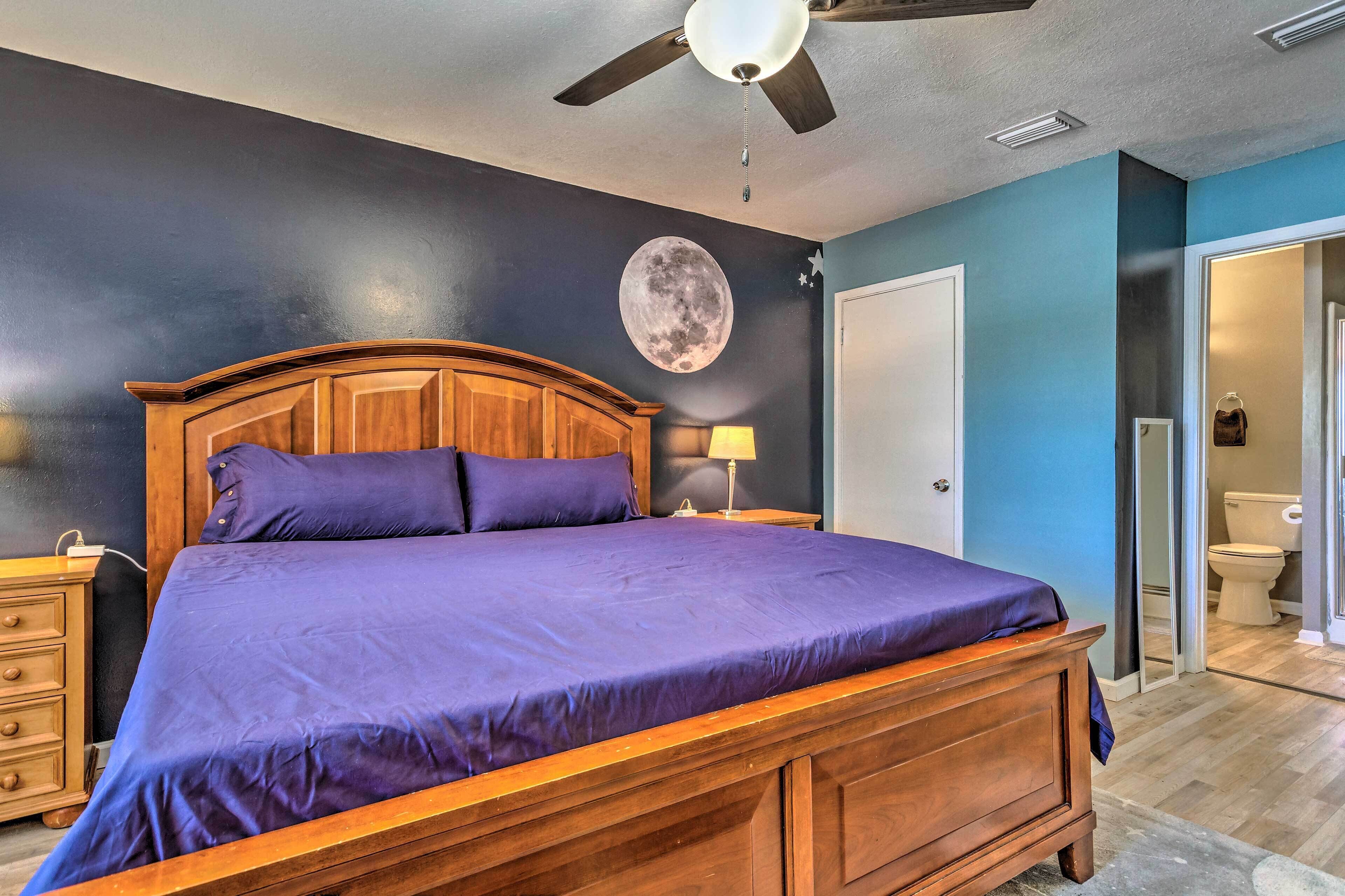 A bedroom with a wooden bed, a ceiling fan, a wall mural of the moon, and a bathroom visible through an open door.