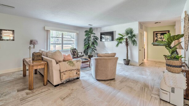A spacious living room with a beige sofa, a round wicker chair, and a small wooden table. There are several potted plants and a large window providing natural light. - Sunny Crystal River Home w/ Screened-in Porch (Crystal River)