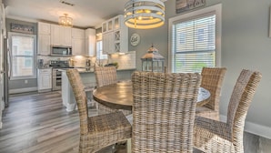 A kitchen with white cabinets, a round dining table, wicker chairs, and a hanging light fixture. - On-site Beach Bar: Sunny Perdido Key Cottage! (Pensacola)