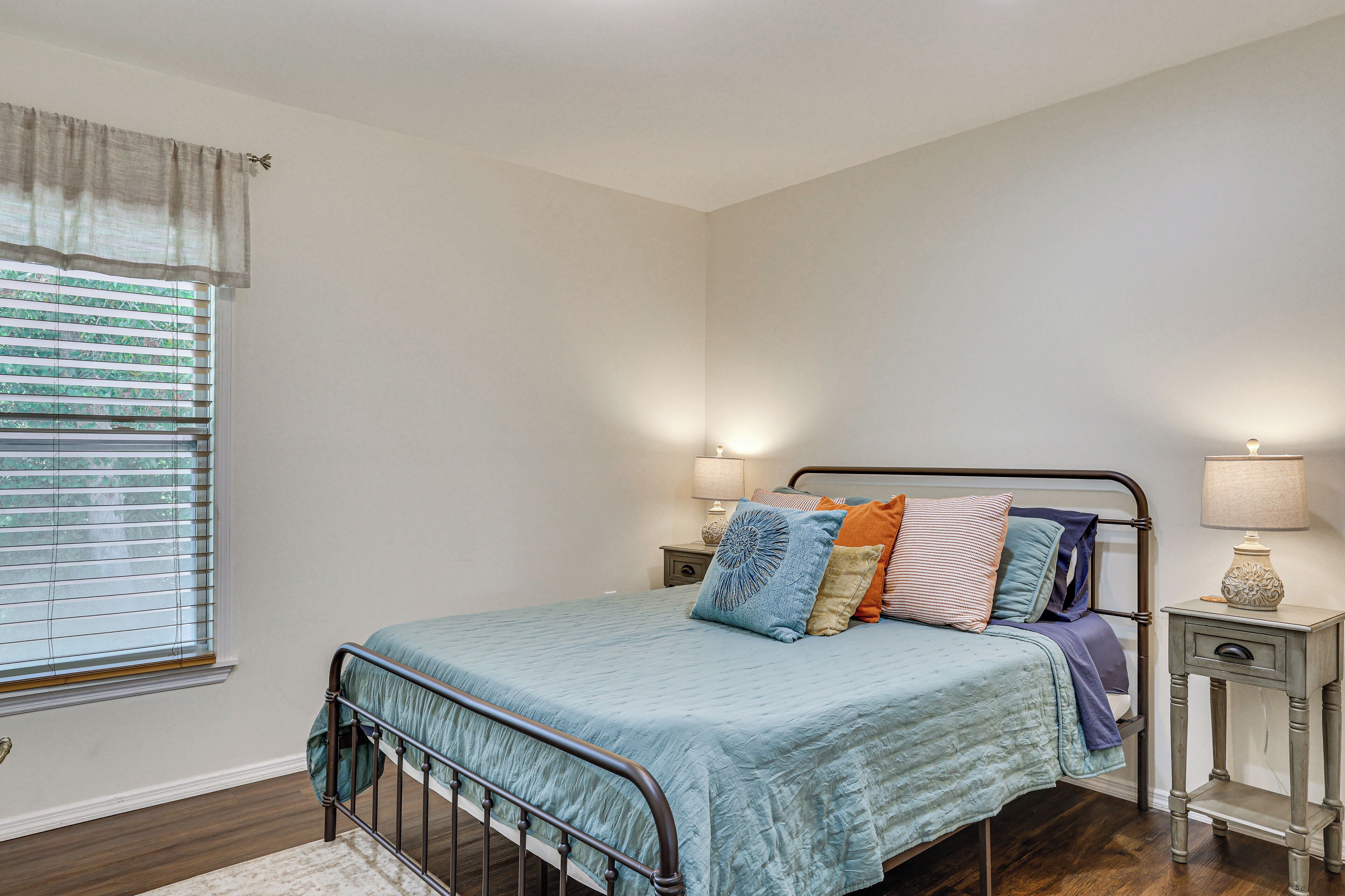 A bedroom with a bed, bedside tables, a window with blinds, and a nightstand with a lamp.