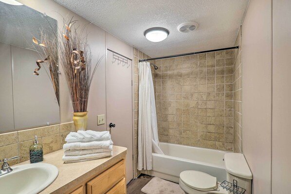 Un baño con lavamanos, inodoro y bañera. Hay un perchero con toallas, un florero con plantas secas y un espejo.