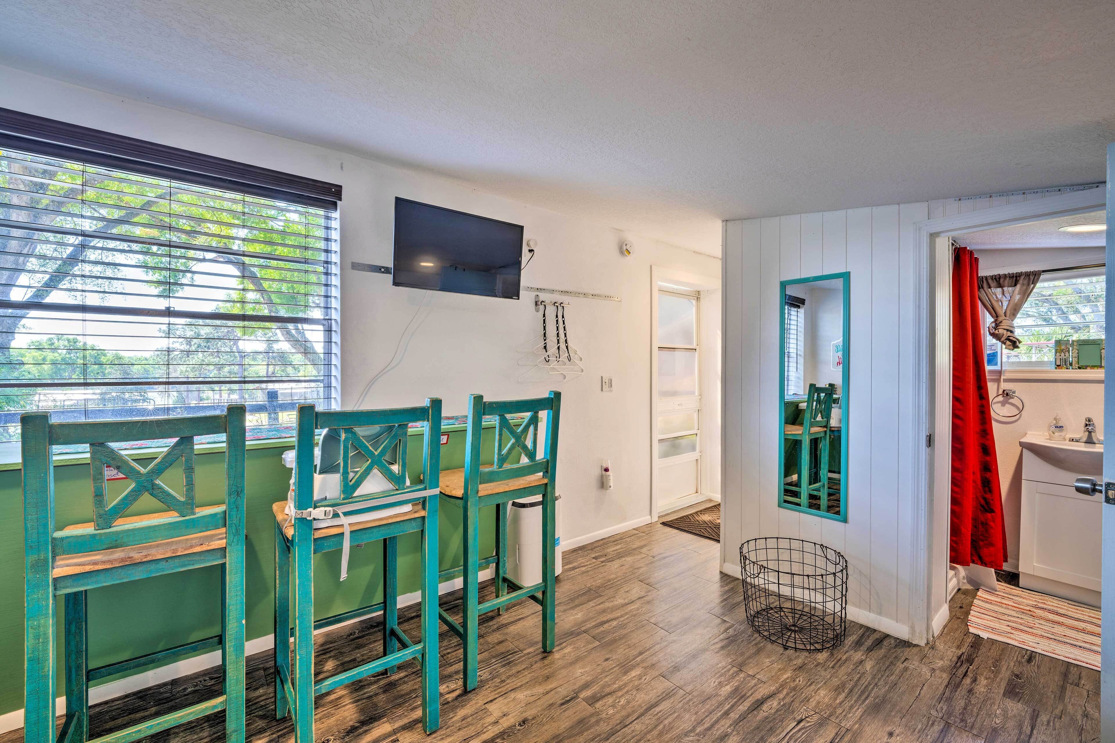 A room with a wooden floor, a green table with chairs, a TV mounted on the wall, and a bathroom visible through an open door.