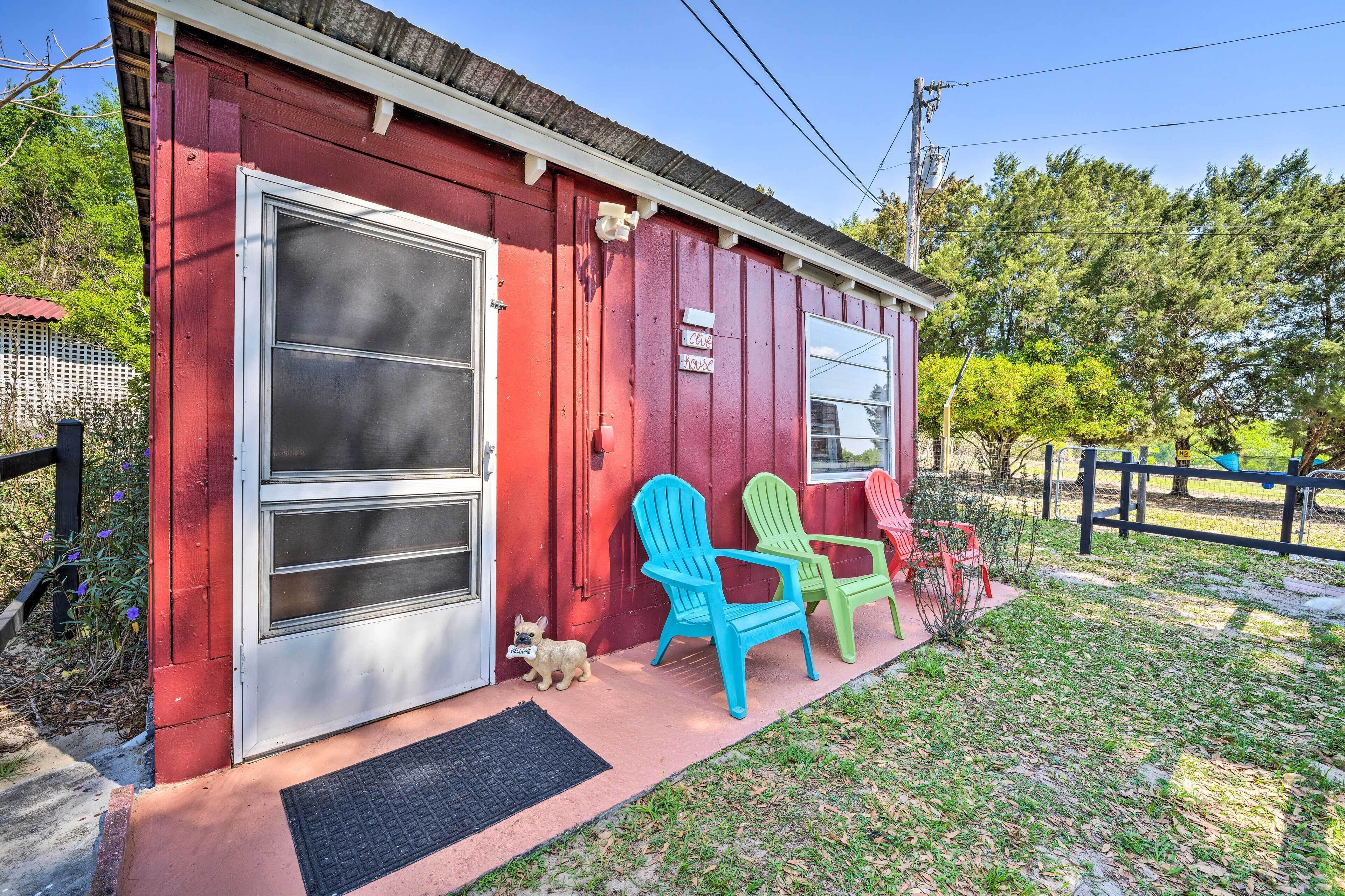 A red building with a large sliding glass door, two Adirondack chairs, and a small dog on the porch.
