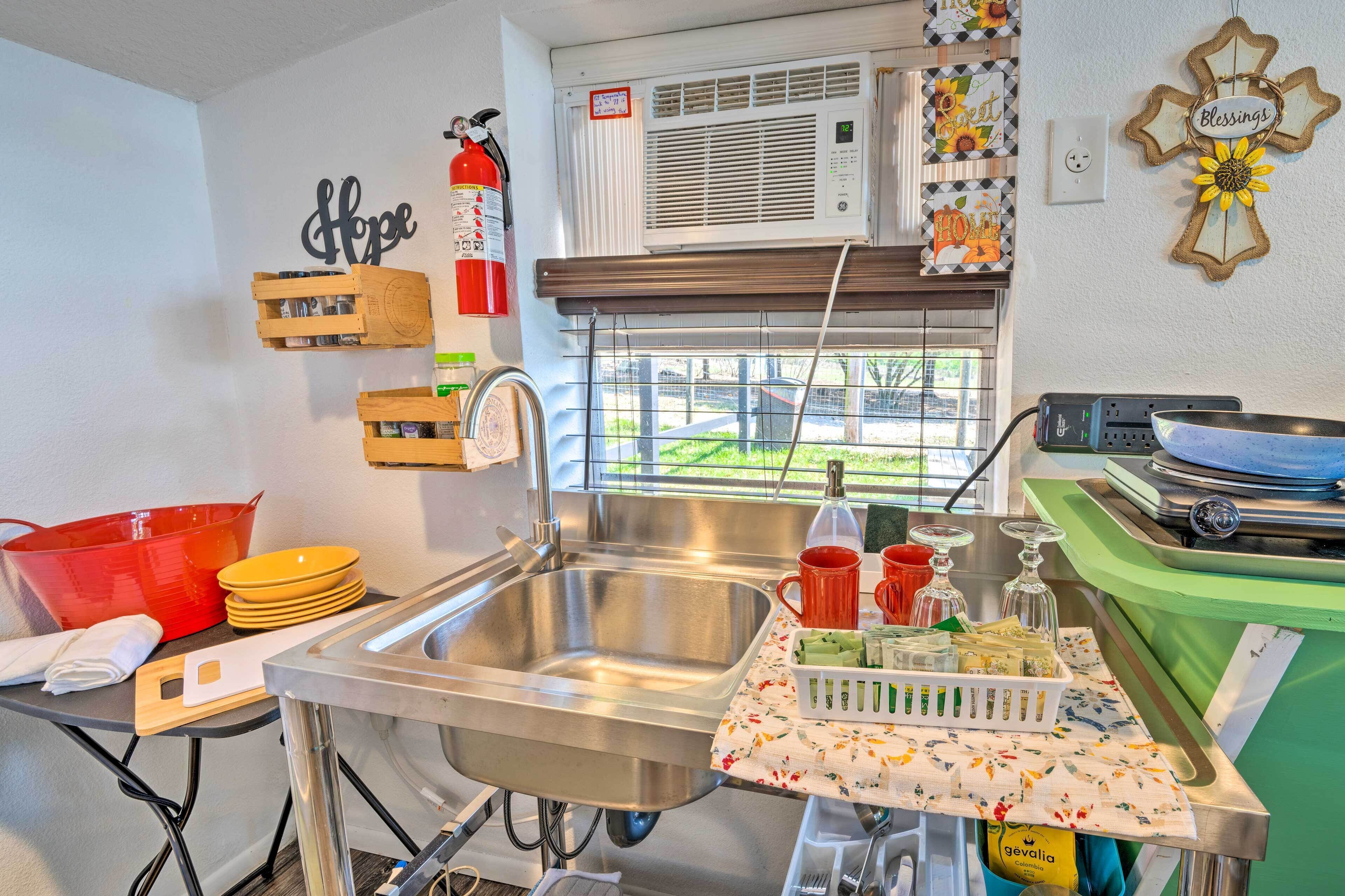 Una cocina con un lavamanos de acero inoxidable, cajas de madera, un cubo rojo y una ventana con vistas a la vegetación.