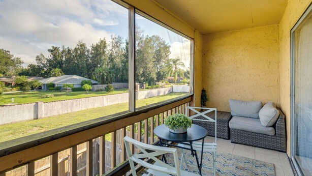 A balcony with a view of a residential area, a wicker sofa, a small table with a plant, and a white chair.