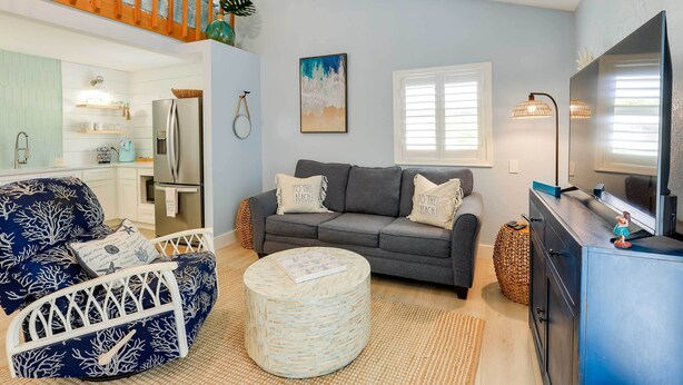 A living room with a grey sofa, a round ottoman, a white armchair, a kitchen area with a fridge, and a staircase. - Tropical Cottage < 2 Mi to Hobe Sound Beach! (Hobe Sound)