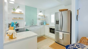 A modern kitchen with white cabinets, a stainless steel refrigerator, and a built-in oven. There are wooden shelves with kitchen items and a small window above the sink. - Tropical Cottage < 2 Mi to Hobe Sound Beach! (Hobe Sound)