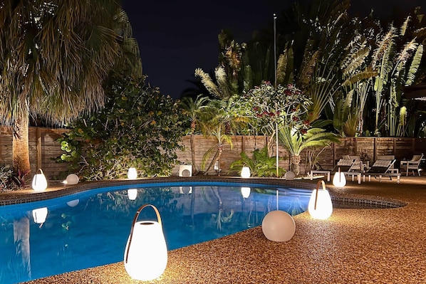 A nighttime pool area with a clear pool, surrounded by palm trees and plants, and illuminated by spherical lights.