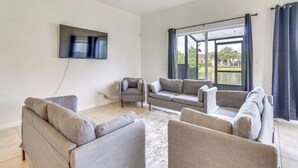 Un salon avec un canapé gris, un petit fauteuil, un téléviseur à écran plat fixé au mur et une grande fenêtre avec des rideaux.