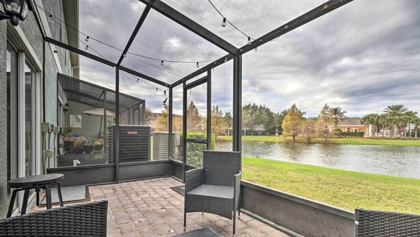 Une terrasse extérieure couverte, aménagée avec des meubles en osier, offrant une vue sur un lac et donnant sur un bâtiment doté d’une porte en verre.