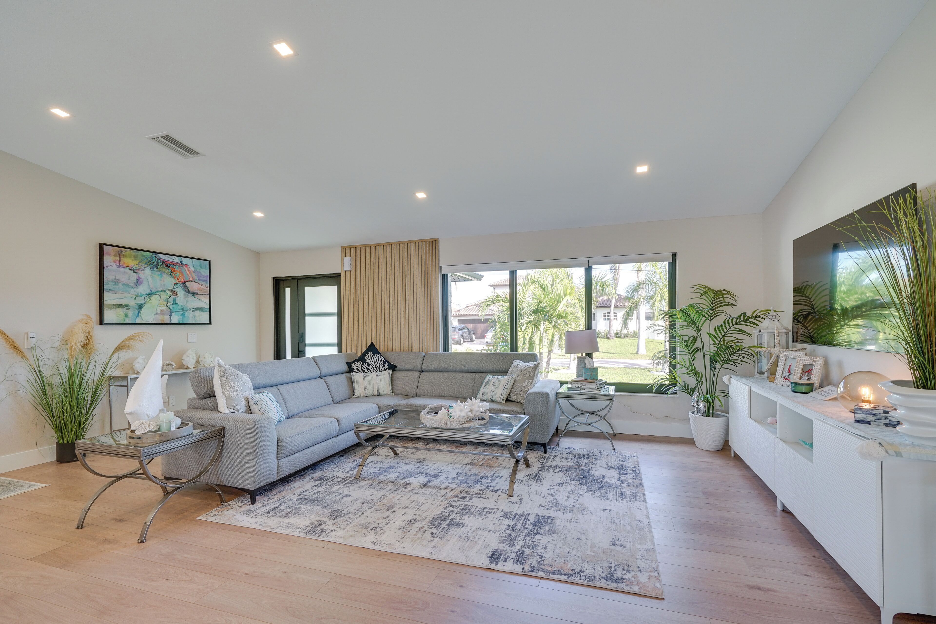 A modern living room with a grey sectional sofa, a glass coffee table, and a large window offering a view of palm trees.