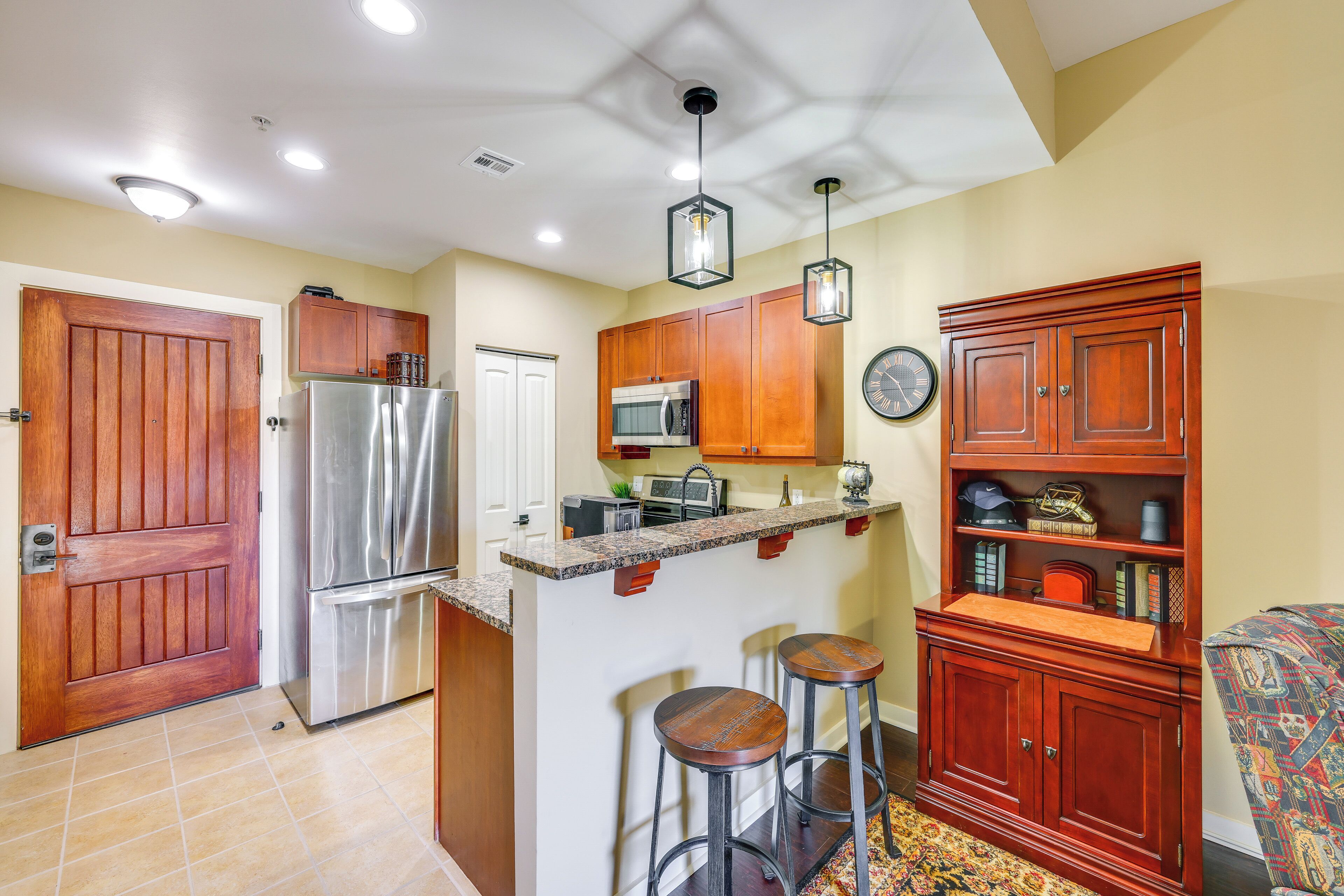 A kitchen with wooden cabinets, a stainless steel refrigerator, a microwave, and a sink. There are two bar stools and a clock on the wall.