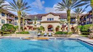 A swimming pool with a fountain, surrounded by palm trees and a building with balconies.
