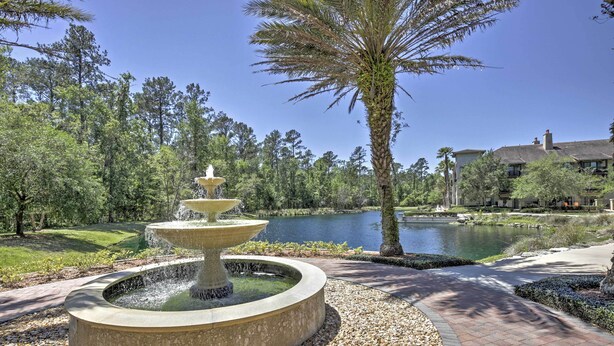 A circular stone fountain with water spouts, surrounded by a pebble path, with a lake and trees in the background.