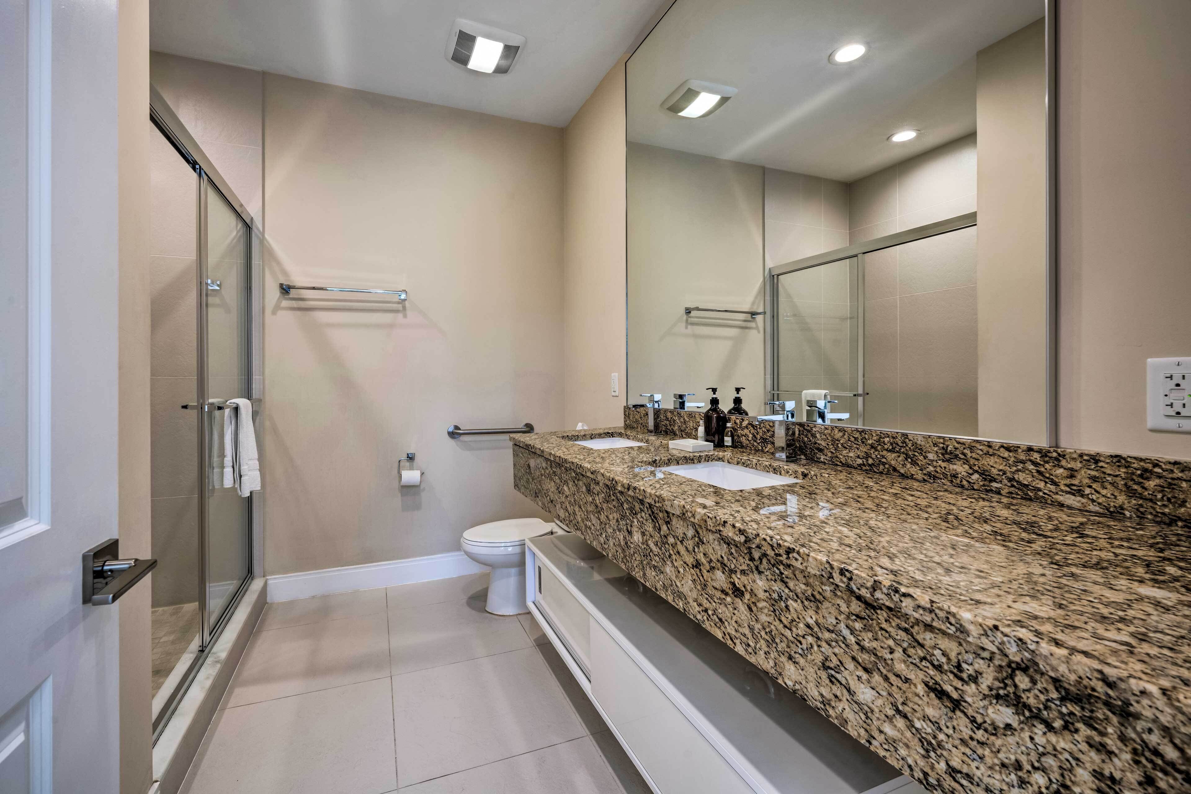 A modern bathroom with a glass shower enclosure, a white toilet, and a granite countertop with two sinks.
