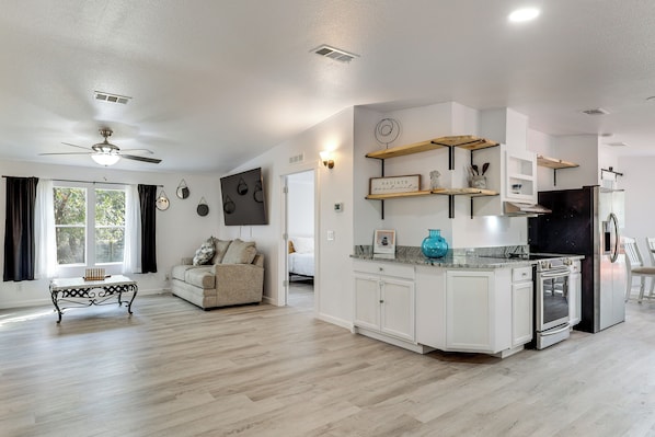 A spacious living room with a ceiling fan, a sofa, and a coffee table. The kitchen features white cabinets, a stove, and a refrigerator.