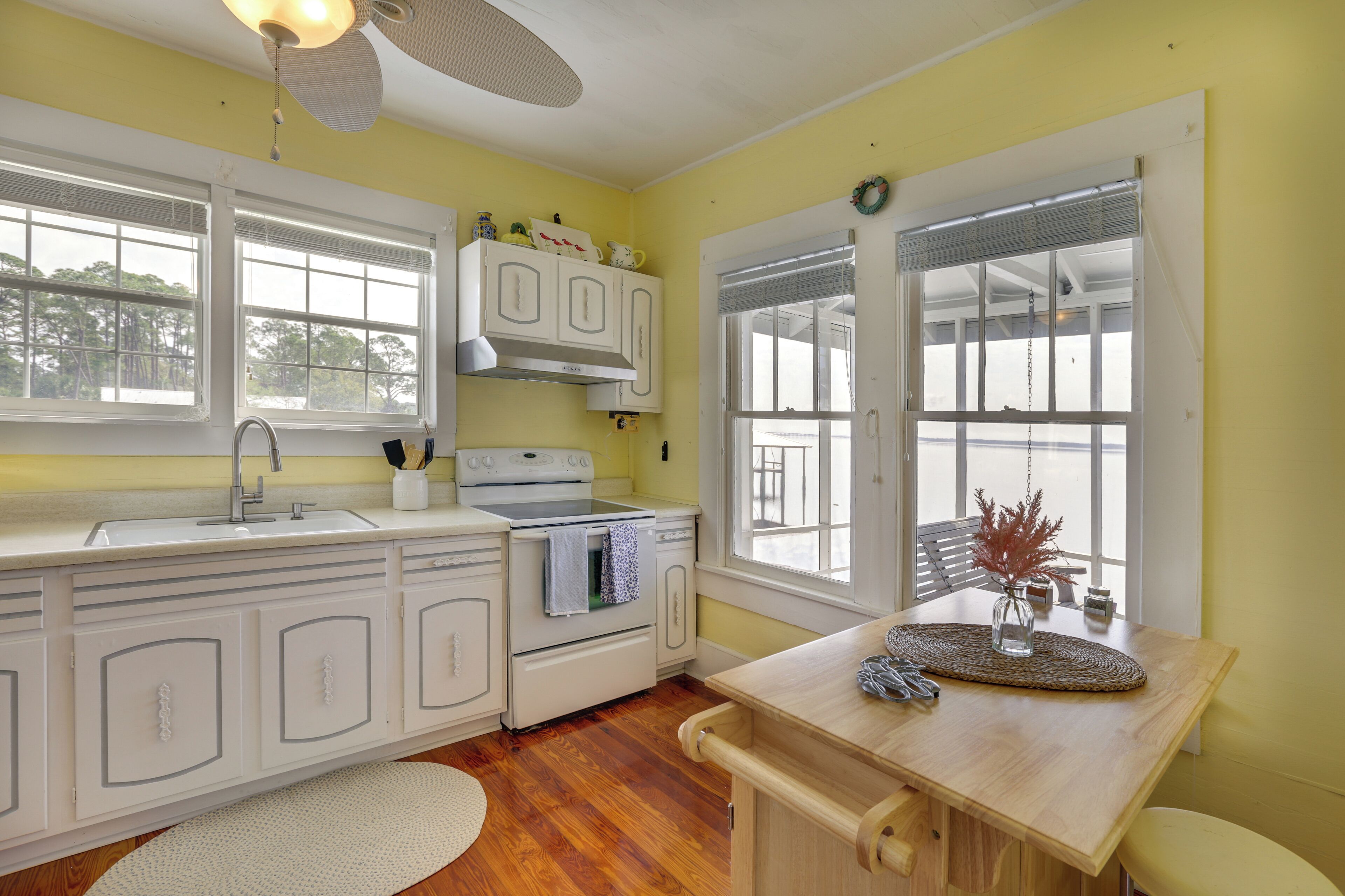 A kitchen with white cabinets, a stove, and a sink. There are windows with blinds and a ceiling fan.