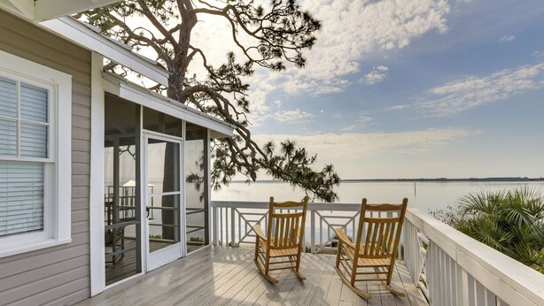 Eine Holzterrasse mit zwei Schaukelstühlen, eine Glastür zu einer Terrasse und Blick auf das Wasser.