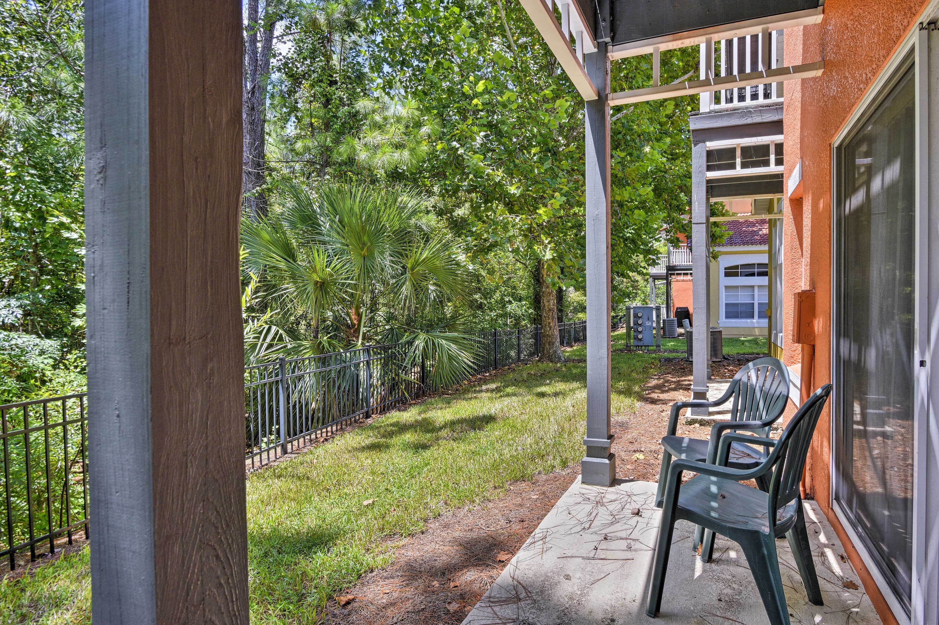 A covered patio with chairs, a concrete pathway, and a fence enclosing a grassy area with trees.