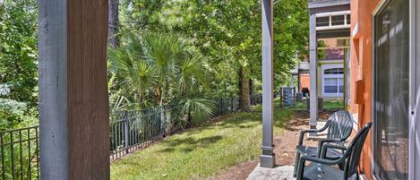A covered patio with chairs, a concrete pathway, and a fence enclosing a grassy area with trees.