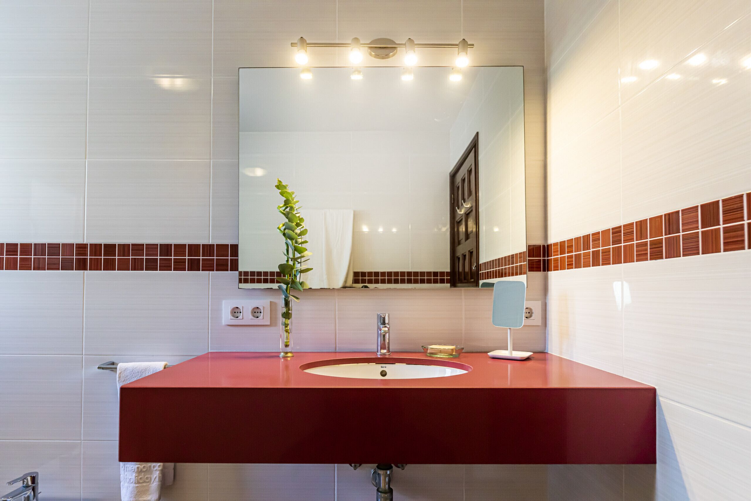 A modern bathroom with a red sink, white tiles, a mirror, and a plant.