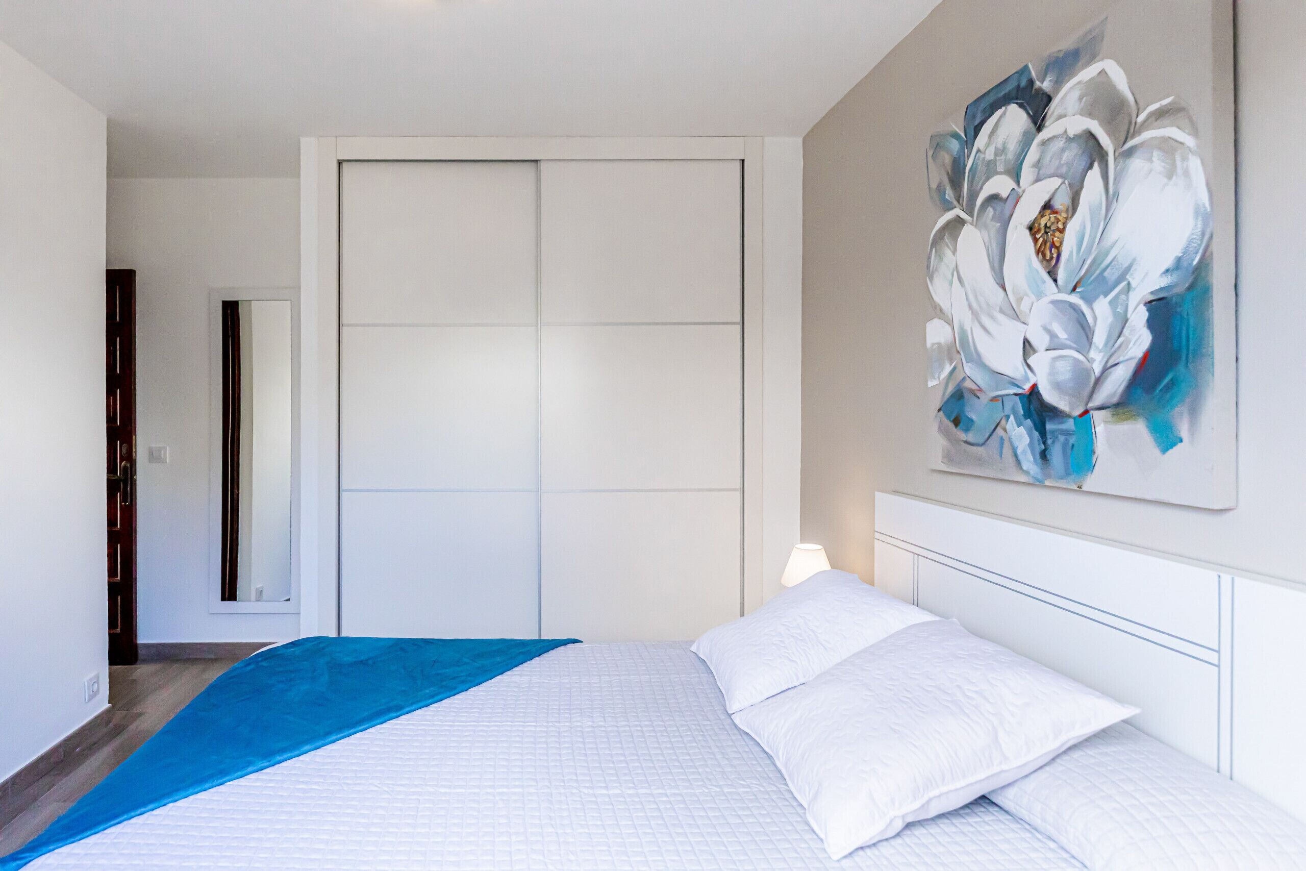 Une chambre à coucher avec un lit, un plaid bleu, un mur blanc orné d’un tableau floral et une armoire.