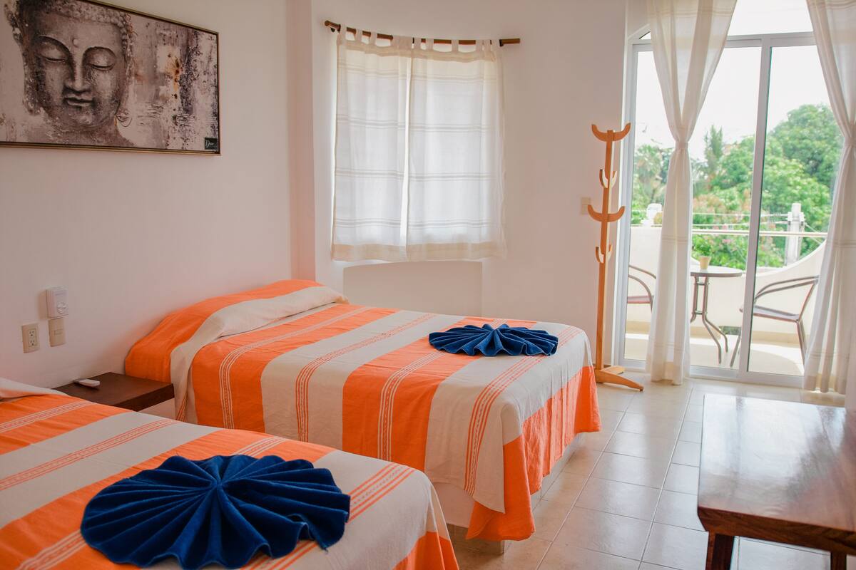 A hotel room with two beds, a window with curtains, a view of greenery outside, and a wooden table.
