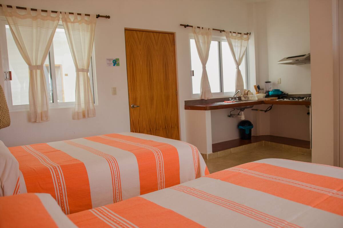 A bedroom with two beds, a wooden door, a window with sheer curtains, and a kitchenette with a stove and sink.