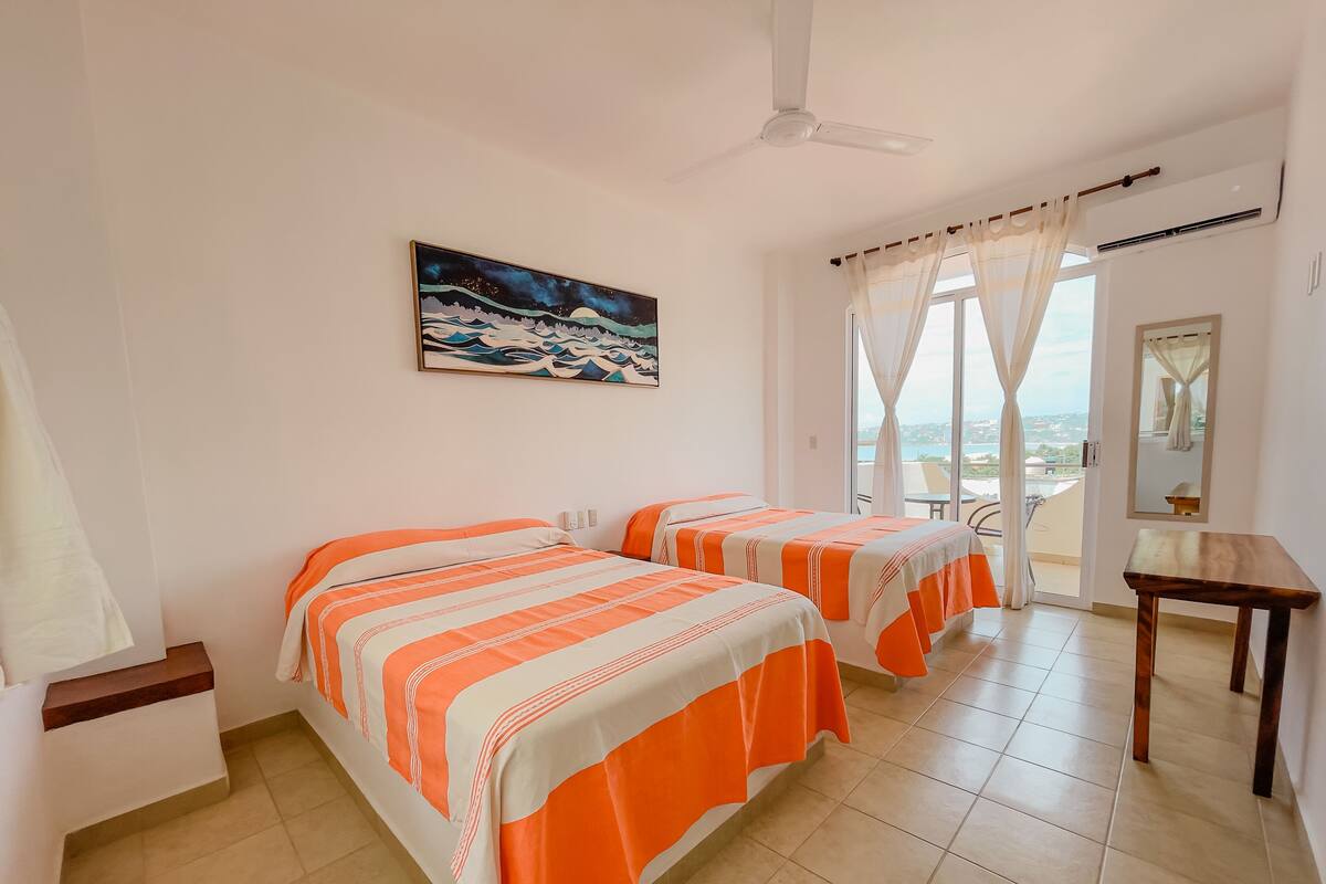 A hotel room with two beds, a ceiling fan, a painting, a window with curtains, and a view of the outside.