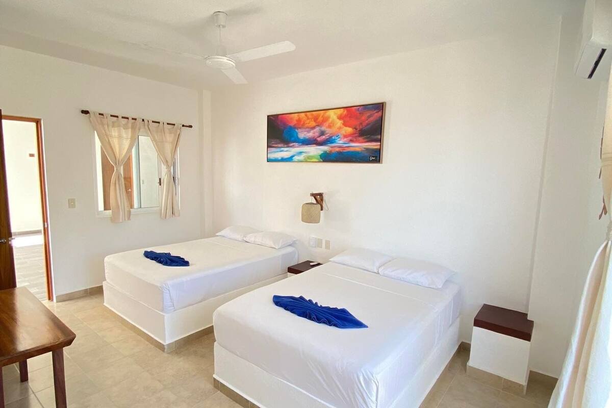A hotel room with two beds, a ceiling fan, a painting, and a small table.