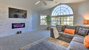 Un salon moderne doté d’une cheminée, d’un ventilateur de plafond, d’une grande fenêtre et d’un canapé d’angle gris agrémenté de coussins orange.
