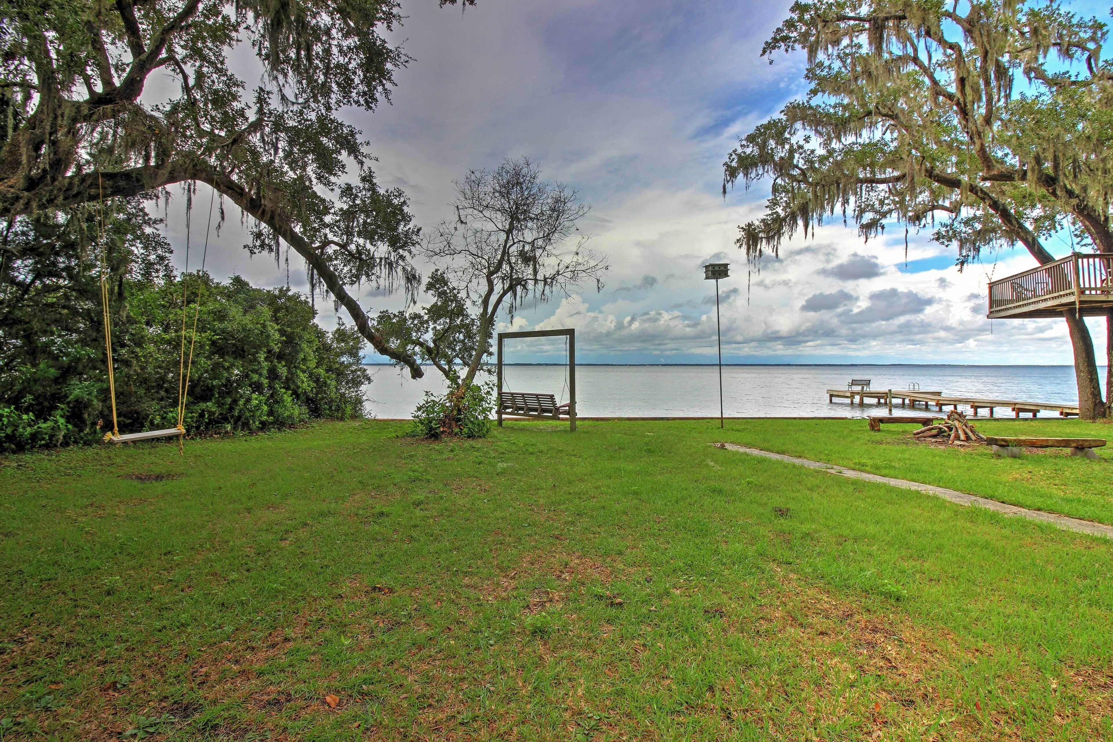 A grassy area with a swing set, a bench, and a wooden deck overlooking a body of water.