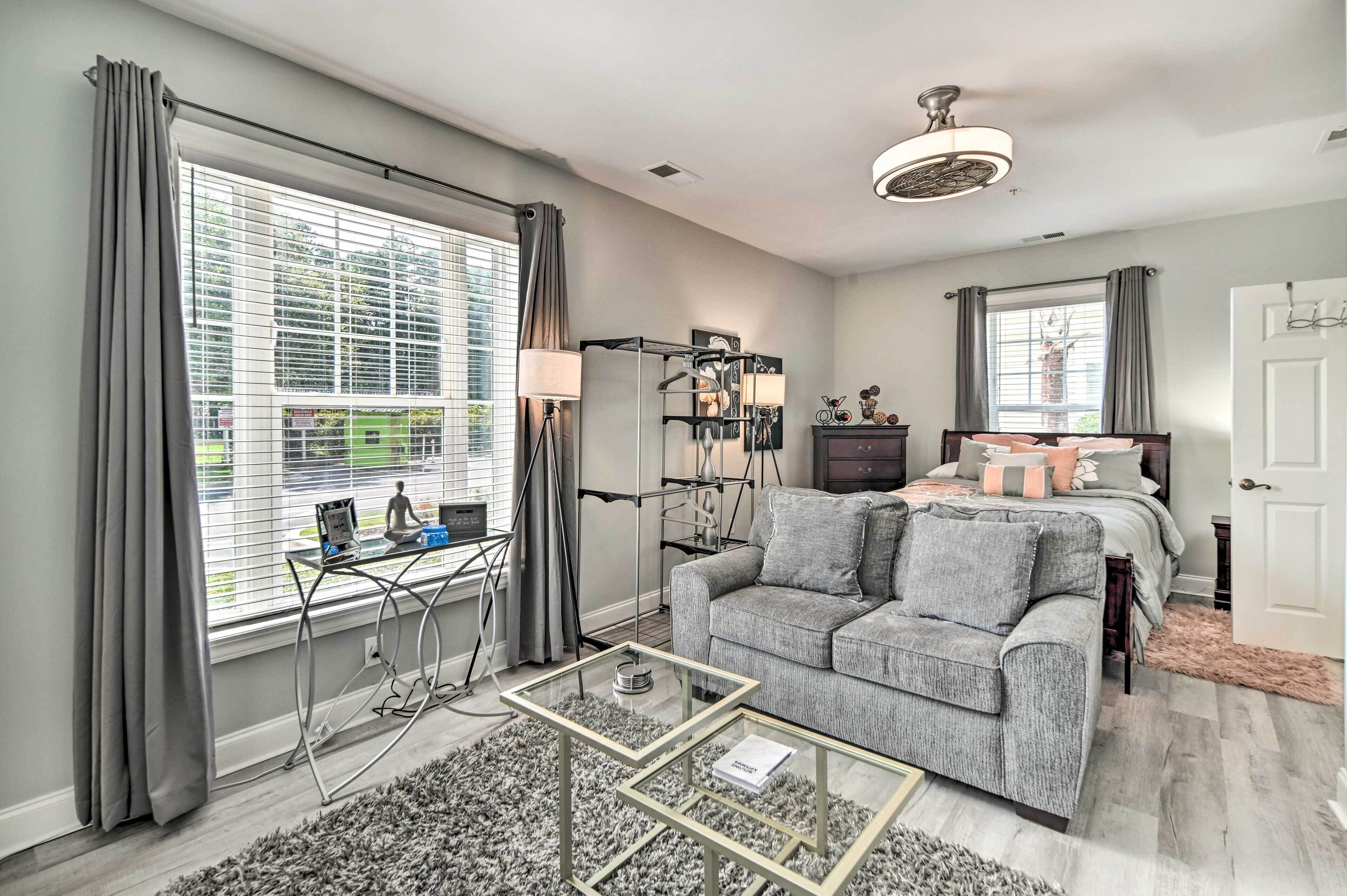 A modern bedroom with a grey sofa, a glass coffee table, a bed with grey bedding, and a large window with blinds.