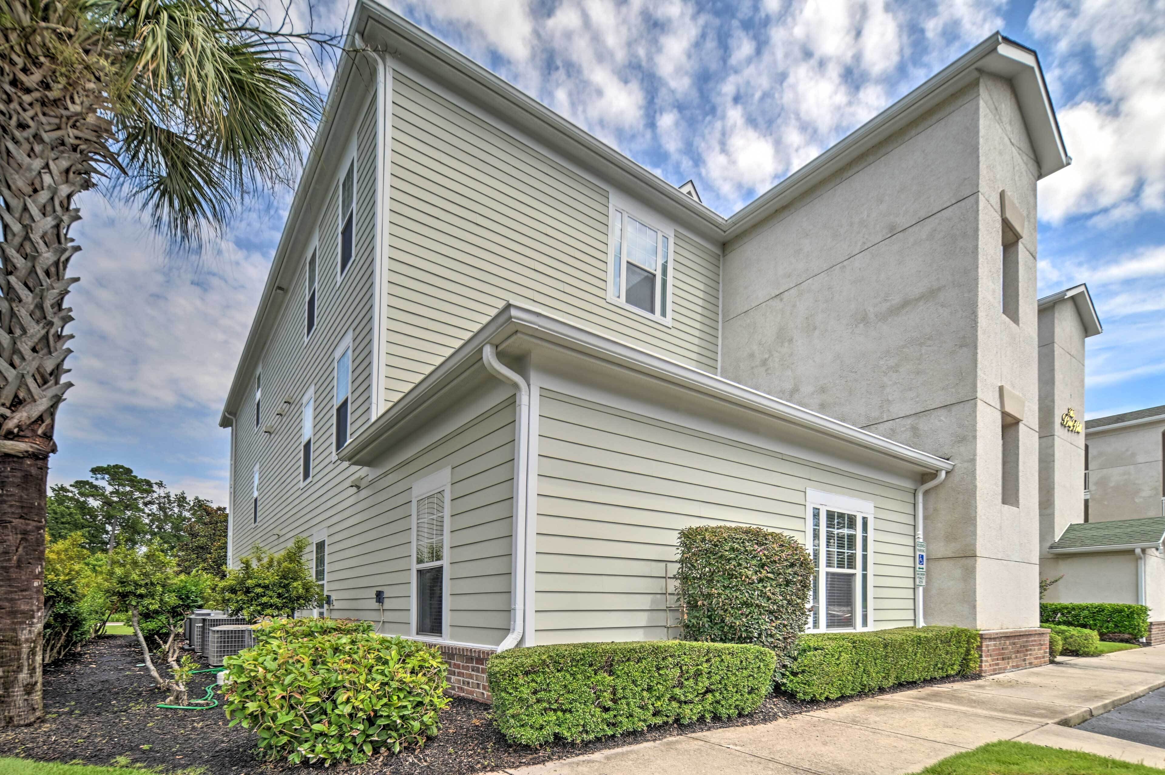A two-story residential building with a symmetrical facade, a small window on the upper floor, and a well-maintained front yard with hedges and a palm tree.