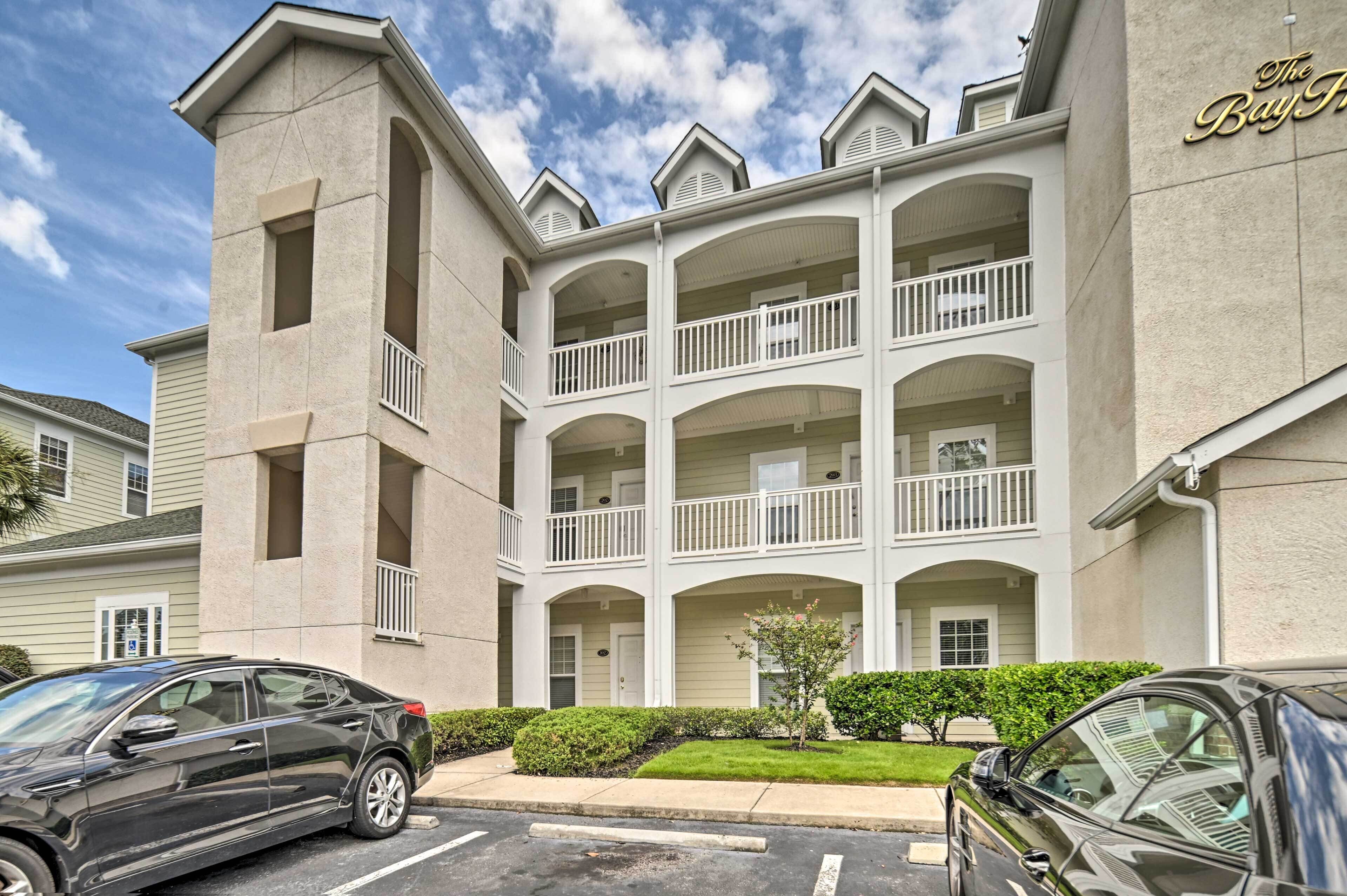 A multi-story hotel with balconies, a parking lot, and a well-maintained lawn.