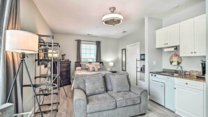 A modern living room with a grey sofa, a metal shelf unit, a lamp, and a kitchen area with white cabinets and a countertop.