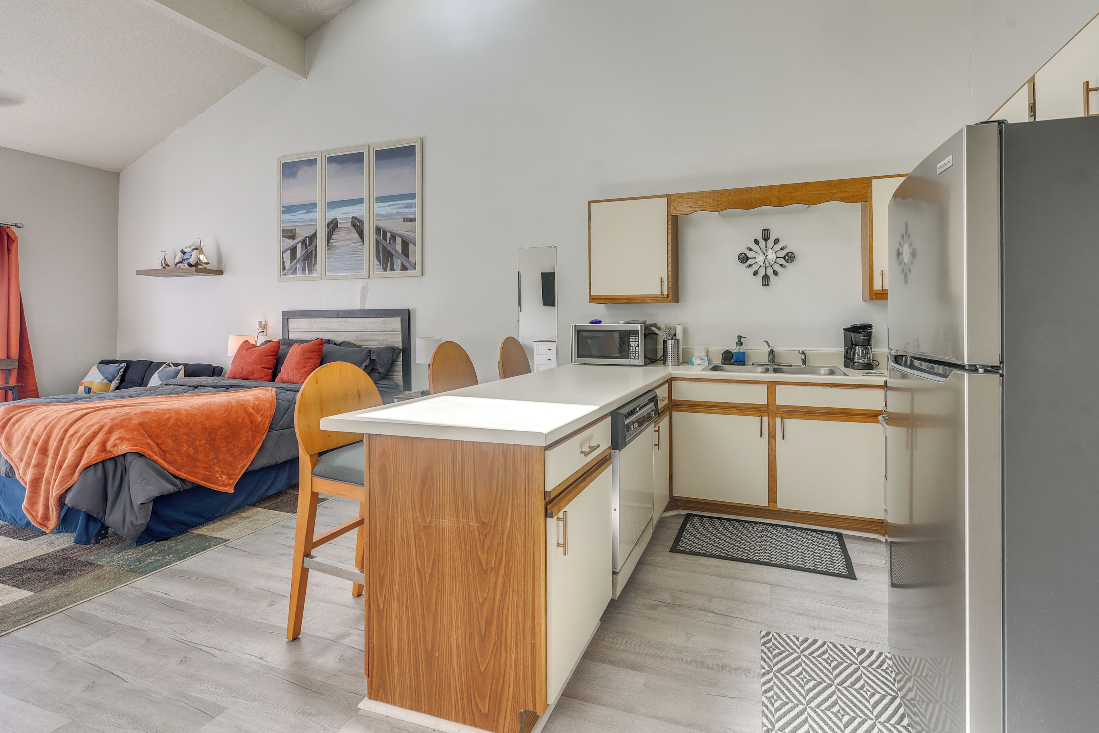A compact kitchen with a microwave, refrigerator, and wooden cabinets. A breakfast bar is adjacent to the kitchen, and a bed with an orange blanket is visible in the background.