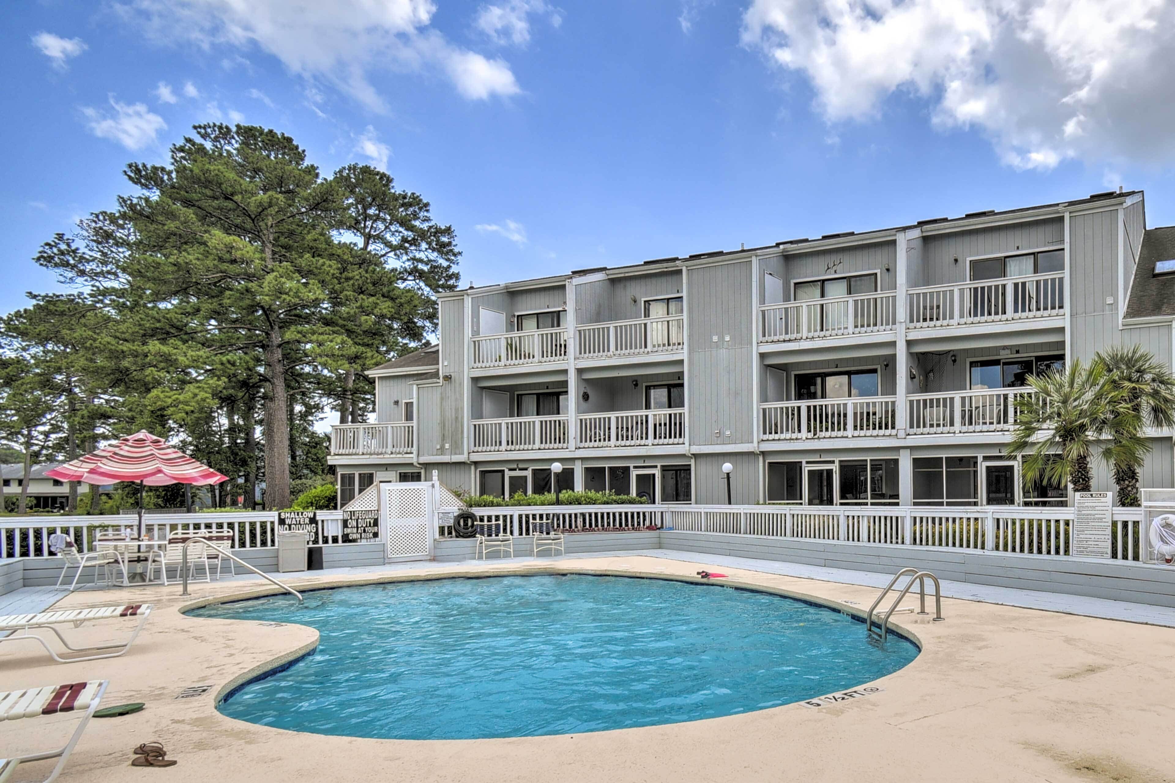 A swimming pool with a curved edge, surrounded by a white fence. There are lounge chairs and a red and white striped umbrella. A multi-story building with balconies and windows is visible in the background.