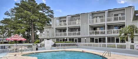 A swimming pool with a curved edge, surrounded by a white fence. There are lounge chairs and a red and white striped umbrella. A multi-story building with balconies and windows is visible in the background.